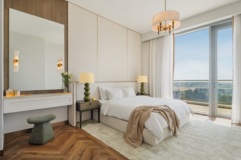 Bedroom with a cream panelled headboard wall, green bubble lamps, a brass chandelier and floor-to-ceiling glass doors to a balcony with an open view