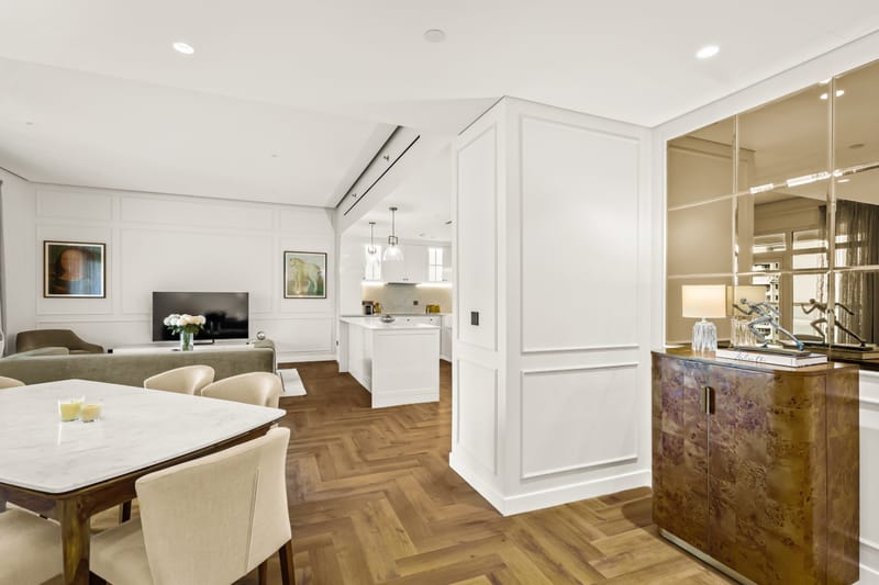 Open-plan living and dining room with a marble dining table, cream chairs, herringbone oak floors and a white kitchen beyond