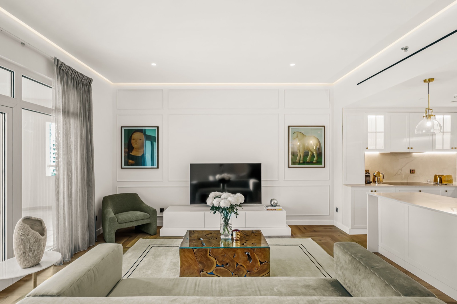 A bright living room in a Laguna Properties residence on Palm Jumeirah, with a sage velvet sofa, a wood and glass coffee table, two framed prints on white panelled walls and an open white kitchen