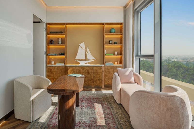 Study lounge with lit burl-wood shelves around a model sailing yacht, a walnut table, blush and cream armchairs and floor-to-ceiling windows over treetops
